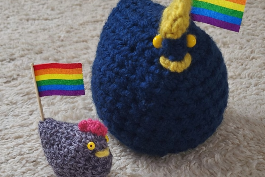 Zwei aus blauem Garn gehäkelte Hühner, die mit einer Regenbogenflagge dekoriert sind.